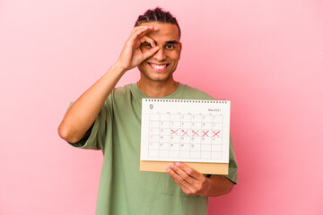 Young venezuelan man holding a calendar isolated on pink background excited keeping ok gesture on eye.