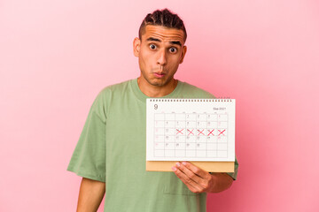 Young venezuelan man holding a calendar isolated on pink background shrugs shoulders and open eyes confused.
