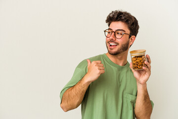 Young caucasian man holding an almond jar isolated on white background points with thumb finger away, laughing and carefree.