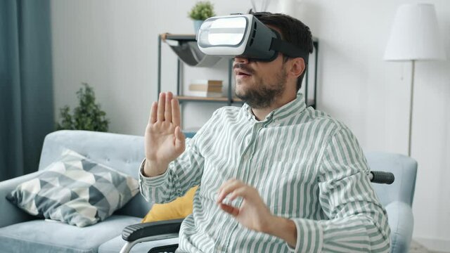 Slow motion of handicapped guy wearing vr glasses moving hands sitting in wheelchair at home alone enjoying trendy device. People and entertainment concept.