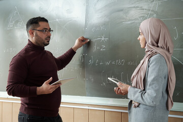 Teacher and student with tablets standing by blackboard at lesson of algebra