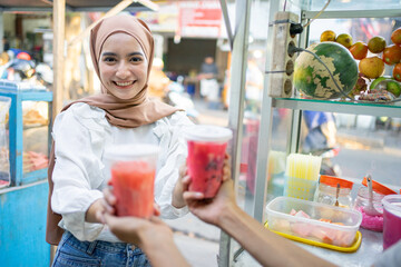 Fototapeta premium a pretty girl in a head scarf smiles at the camera as she picks up two cups of fruit ice from the seller beside the cart