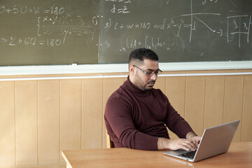 Serious teacher typing by desk against blackboard in classroom