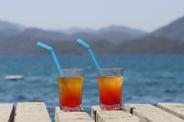 Two glasses with cocktail on a wooden pier on sea and mountains background. Summer vacation on a beach