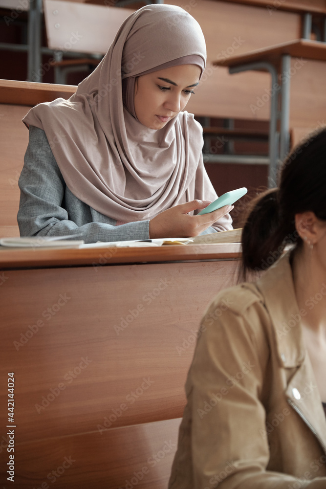 Poster girl in hijab scrolling in smartphone by desk during lecture