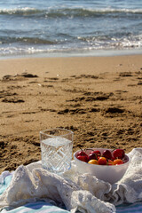 Picnic on the beach, bowl with sweet cherry, glass of water on the sand. Calm blue sea on background. Summer day photo. 