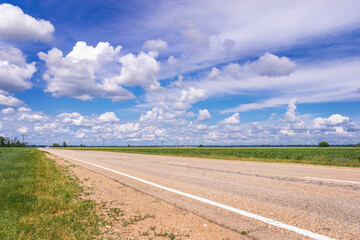 Summer landscape. View of an asphalt road through a field with green grass. Highway through a field against a blue sky with white cumulus clouds.