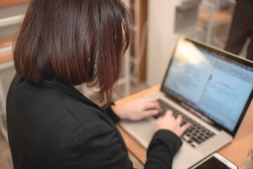 An anonymous woman of medium length hair composes a document on a laptop while at coworking space. Remote work at a cafe.