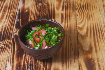 Vegetable salad in a clay bowl on a wooden table, sliced vegetables tomatoes, cucumber, dill, onion, parsley, radish sprinkled with salt and vegetable oil