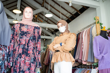Fototapeta na wymiar asian woman in a hijab wearing a mask and crossing hands stands inside a boutique shop