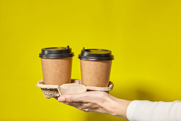 Hands holding two cups brown paper with black lid. Two coffee special offer or promo. Hands holding two cups on yellow screen background. Tea or Coffee to go. Brown paper cup with black lid.