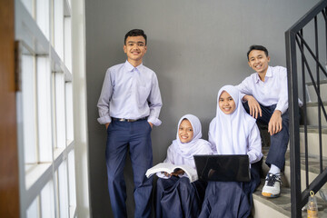 four teenagers in junior high school uniforms smile at the camera while holding a laptop and a book while sitting on the stairs