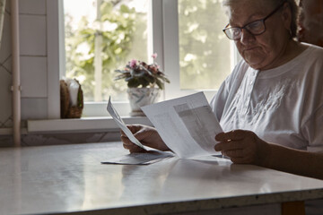 Elderly woman sitting at table in kitchen at home holds domestic bills, feels concerned forgot to pay or debt formed. Middle aged 80s retired woman
