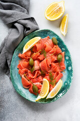 Slices of salted salmon with capers, lemon and dill on a ceramic plate on a gray concrete background. Top view
