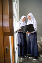 two teenagers wearing headscarves in school uniforms carrying a laptop stand by the window