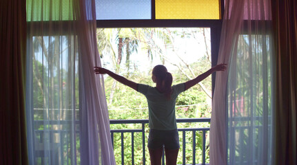 Fototapeta na wymiar Lovely woman opening colorful curtains and meeting sunrise. View from balcony to jungle.