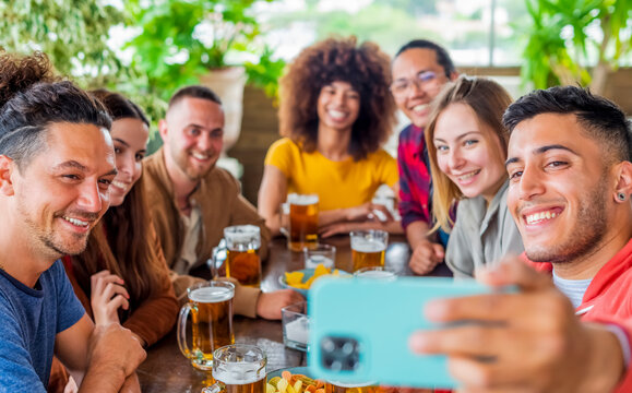 diverse group of friends making a selfie celebrating happy hour with beers and food in a bar restaurant. multiethnic people having fun in a party together smiling at the camera. self portrait concept