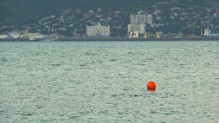 Orange buoy on the water. Ripples in the water. Water marking. Restriction of movement. Signal float. Mountains on the shore. Settlement in the background