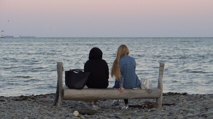 Two girls are sitting on a bench. Without a face. Rocky seashore. Calm water. Evening. Communication of people. Horizon. Pink sky. Ships at sea.