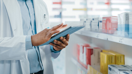 Pharmacy: Portrait of Professional Black Pharmacist Uses Digital Tablet Computer, Checks Inventory of Medicine, Drugs, Vitamins, Health Care Products. Druggist in Drugstore Store. Focus on Hands