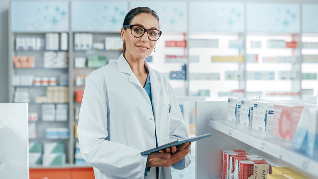 Pharmacy: Portrait of Professional Beautiful Caucasian Female Pharmacist Uses Digital Tablet Computer, Checks Medicine Inventory, Looks at Camera, Smiles Charmingly. Drugstore Health Care Products - Powered by Adobe