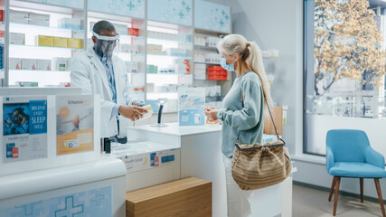 Obraz premium Pharmacy Drugstore Checkout Counter: Professional Black Pharmacist Wearing Face Shield Sells Medicine to Senior Female Customer, who is wearing Face Mask, Use Contactless Payment. Coronavirus Safety