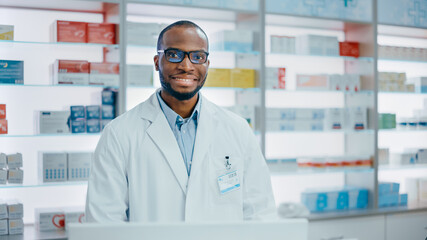 Pharmacy Counter: Portrait of Handsome Professional Black Male Pharmacist Working on Computer, Looks at Camera Smiling Charmingly. Drugstore Specialist, Shelves Health Care Products, Medicine Behind