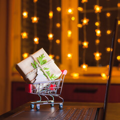 Miniature shopping cart with decorated gift box standing on laptop. Concept of on-line shopping
