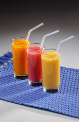 Three glasses of fruit juice with plastic straws over blue placemat on gray gradient background.