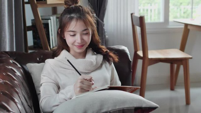Happy Asian teenage woman sit and write note or design draw a picture in notebook. in living room at home. Write memo of planning your work strategy on paper. work stay at home.