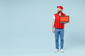 Obraz premium Full size body length delivery guy employee man in red cap white T-shirt vest uniform workwear work dealer courier hold use laptop pc isolated on pastel blue color background studio Service concept