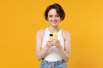 Young smiling happy friendly woman 20s with bob haircut wearing white tank top shirt using mobile cell phone chat online browsing surfing internet isolated on yellow color background studio portrait.