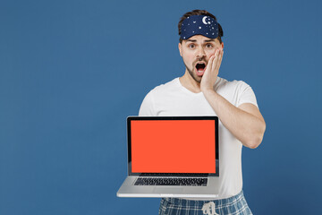 Young shocked sad man in pajama sleep mask rest relax at home hold using laptop pc computer with blank screen workspace area hold face isolated on dark blue background Bad mood night bedtime concept.