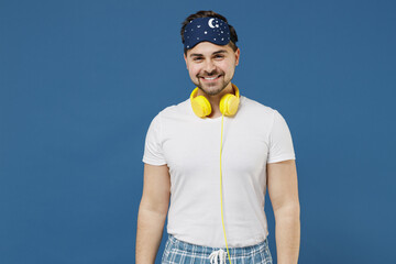 Young smiling fun caucasian happy man 20s in pajamas jam sleep mask headphones resting relaxing at home listen to music audiobook isolated on dark blue sky background Good mood night bedtime concept