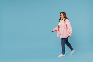 Full length side view young fun smiling redhead chubby overweight woman 30s wearing in pink shirt jeans casual clothes walking going looking aside isolated on pastel blue background studio portrait