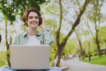 Young smiling student fun freelancer woman in green jacket jeans sit on bench in spring park outdoors rest use laptop pc computer wi-fi internet work online look camera People urban lifestyle concept.