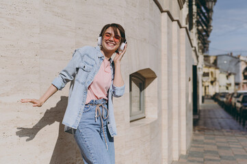 Young excited fun overjoyed caucasian woman 20s wearing jeans clothes headphones eyeglasses listening to music dancing outdoors on city street near building wall. People urban youth lifestyle concept