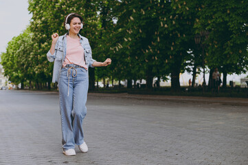 Full length young fun excited caucasian student woman 20s wearing jeans clothes headphones listening to music walking strolling down green park alley outdoors, dancing. People urban lifestyle concept