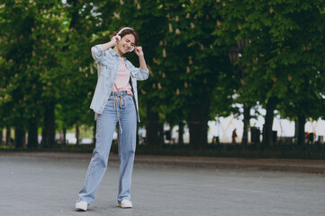 Full length young fun happy student excited caucasian woman 20s wear jeans clothes headphones listen to music walking strolling down green park alley outdoors dancing People urban lifestyle concept