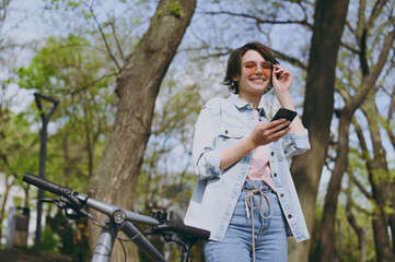 Fototapeta na wymiar Bottom view young smiling caucasian fun woman in jeans clothes stand near bicycle bike on sidewalk in city spring park outdoors use mobile cell phone look aside. People active urban lifestyle concept.