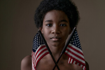 Young dark-skinned boy holding American flags and looking at camera on black background