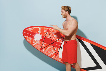 Side view young sexy sporty man 20s wear red shorts swimsuit hold orange sup board point finger...