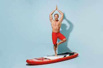 Full length young sexy sport man 20s wear red shorts swimsuit relax stand in yoga pose on orange sup board surfing meditate isolated on pastel blue background Summer vacation sea rest sun tan concept.