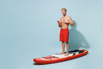 Full length young sexy man 20s wear red shorts swimsuit stand on orange sup board point finger on...