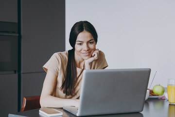Young happy freelenacer copywriter housewife woman 20s in beige shirt eat breakfast using laptop pc...