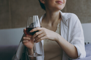 Close up cropped photo shot young woman in white clothes plaid sit on sofa indoors apartment hold glass grink red wine prop up face look aside. Rest on weekends leisure quarantine stay home concept.