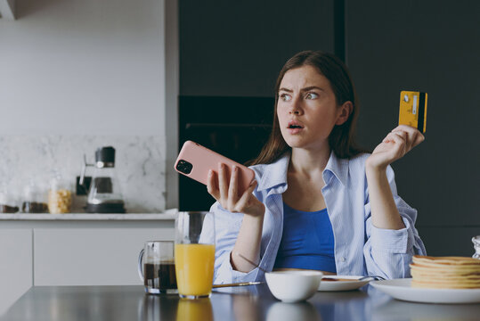 Young Puzzled Sad Housewife Woman In Casual Clothes Eat Breakfast Use Mobile Cell Phone Credit Bank Card Shop Online Order Delivery Cook Food In Light Kitchen At Home Healthy Diet Lifestyle Concept.