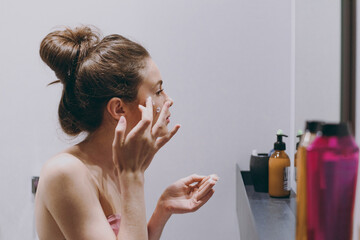Fototapeta premium Side view half naked topless young woman 20s applying moisturizer, anti-aging facial cream standing in front of mirror in bathroom do morning routine Skin care healthcare cosmetic procedures concept.
