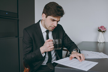 Young serious smart pensive employee business man corporate lawyer 20s wearing formal black suit shirt tie drink coffee breakfast read paper documents sit at table in kitchen People lifestyle concept