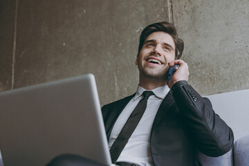 Bottom view smiling young employee business man corporate lawyer in black suit shirt tie sitting on grey sofa at home indoors use laptop pc computer talk mobile cell phone People lifestyle concept